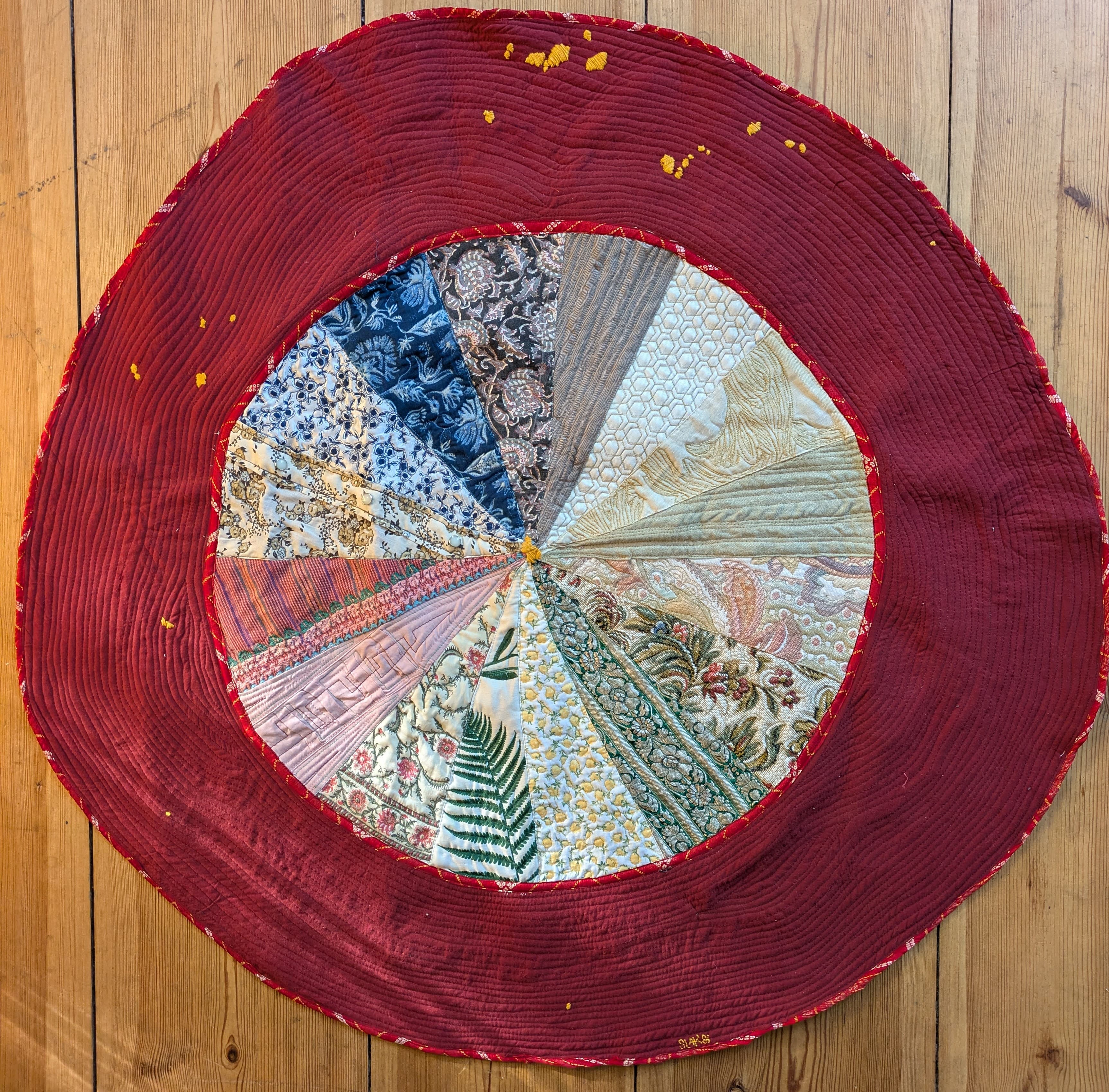 a round patchwork quilt of many fabrics around a red quilted border, stitched with small yellow marks.