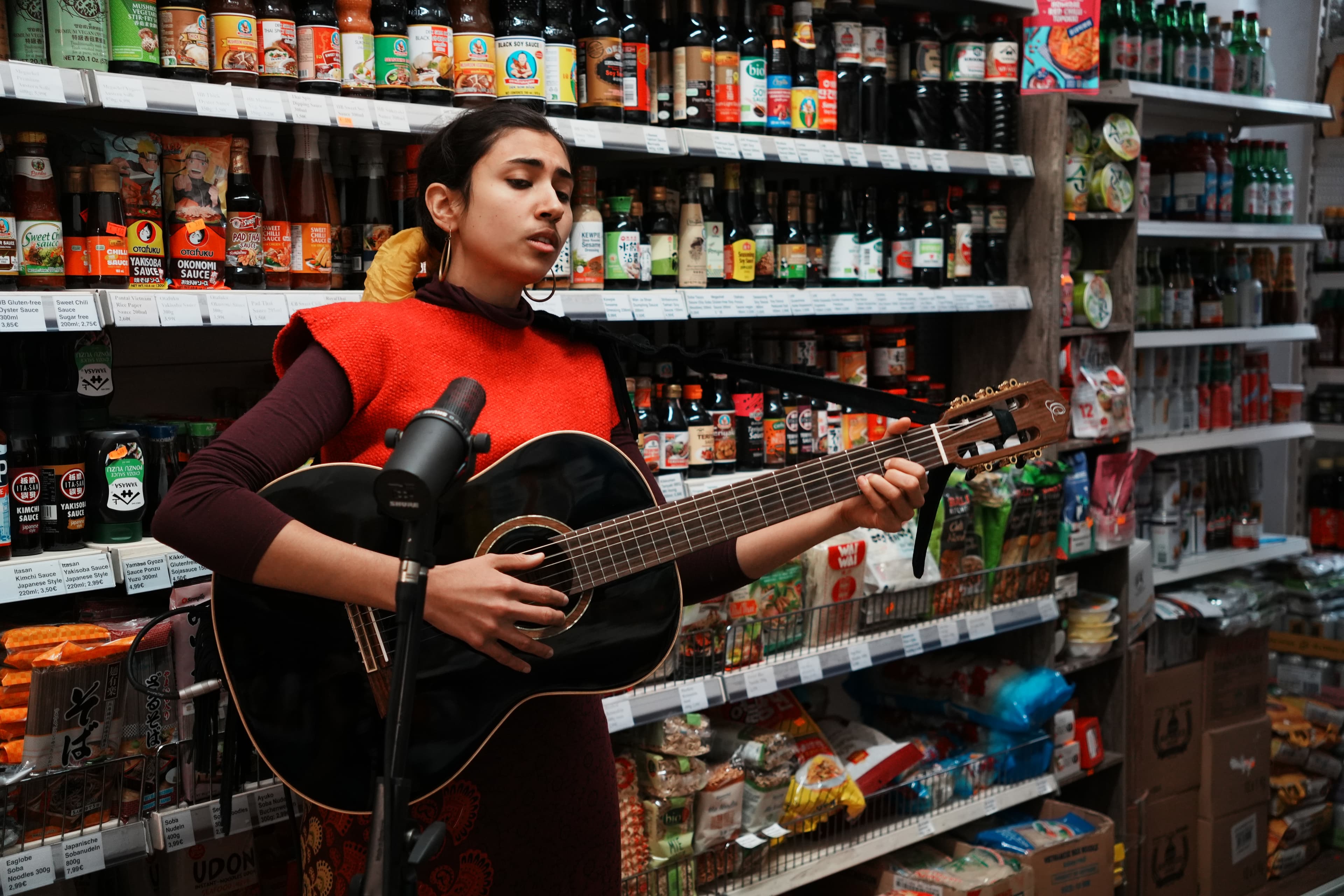 saba lou singing and playing guitar in front of a microphone, in a shop lined with shelves.